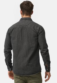 Camisa de mezclilla negra con mangas largas y cuello abotonado. Presenta detalles de costura y aberturas laterales en los puños.
