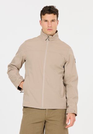 Man draagt een beige jas met rits en hoge kraag en kaki broek, staat met één hand in zijn zak tegen een witte achtergrond.