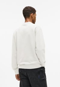 Rövid, göndör hajú férfi fehér hosszú ujjú pulóverben és fekete farmernadrágban, háttal állva egy fehér háttér előtt.