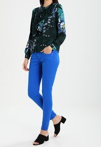 Blouse florale à volants en vert foncé et bleu, associée à un pantalon slim bleu vif. Des sandales à block heels ouvertes noires complètent le look.