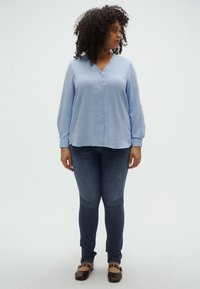 Ανοιχτό μπλε πουκάμισο με υφή και κουμπιά μπροστά, με μακριά μανίκια, συνδυασμένο με σκούρο τζιν και καφέ φλατ παπούτσια. Το πουκάμισο έχει V-λαιμόκοψη.