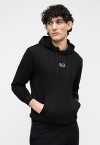 Juodas džemperis su kapišonu, pagamintas iš minkštos medžiagos, su kengūros kišene, raišteliais prie kapišono ir maža auksine logotipu ant krūtinės. Klasikinio kirpimo.