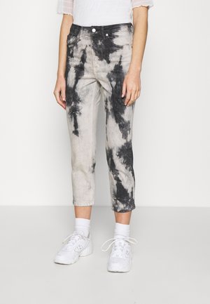 Personne portant un jean court tie-dye noir et gris, des chaussettes blanches, des baskets blanches et un haut blanc à manches courtes, debout sur un fond neutre.