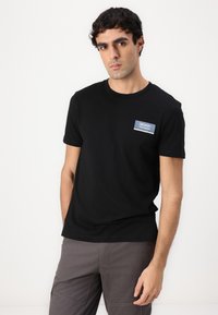 Sort bomulds T-shirt med rund halsudskæring og korte ærmer. Har et blåt denim-inspireret mærke på venstre bryst. Simpelt design, ingen mønstre.