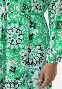 Groene jurk met een patroon van bloemen in tinten groen, zwart en blauw. Gemaakt van lichte, gestructureerde stof met een tailleriem.