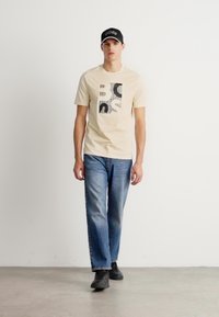 Young man walking, wearing beige graphic t-shirt, blue jeans, black sneakers, and black cap against plain white background.