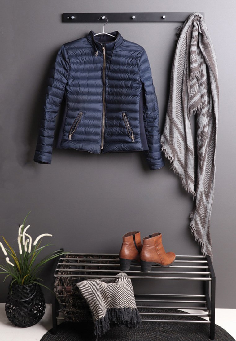 Doudoune bleue sur un portant mural, écharpe beige à motifs suspendue à côté, bottines brunes sur un porte-chaussures en métal, plante dans un pot noir à proximité.