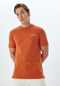 Kortærmet orange bomulds t-shirt med rund hals og hvid broderet tekst på venstre bryst. Enkelt design, blød tekstur.