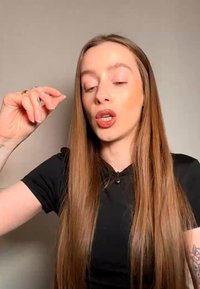 Femme aux longs cheveux raides et chemise noire faisant un signe de la main levée et la bouche légèrement ouverte sur fond uni.