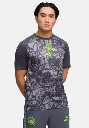 Uomo che indossa una maglietta sportiva grigia del Manchester City e pantaloncini abbinati con loghi verde neon, in piedi davanti a uno sfondo semplice.