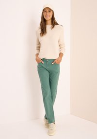 Suéter de punto color beige claro, pantalones verdes con bolsillos y zapatillas de color claro. El modelo posa con las manos en los bolsillos frente a un fondo neutro.