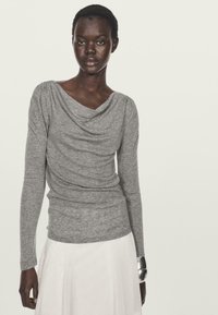 Maglia grigia a maniche lunghe con scollatura drappeggiata, texture morbida e design aderente. Abbinata a una gonna bianca e a un bracciale oversize in argento.