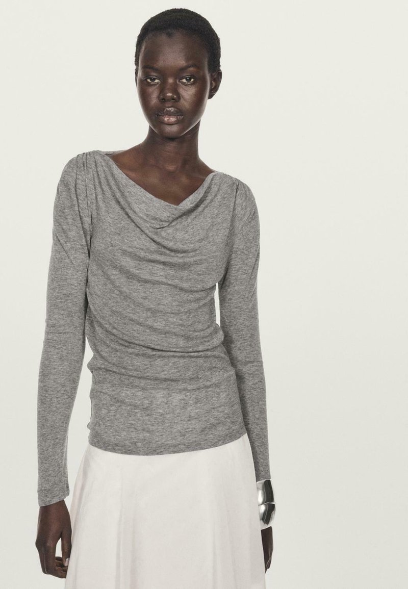 Maglia grigia a maniche lunghe con scollatura drappeggiata, texture morbida e design aderente. Abbinata a una gonna bianca e a un bracciale oversize in argento.