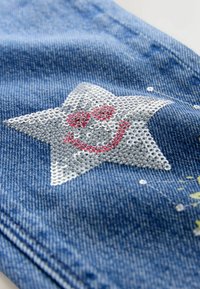 Džínová tkanina s designem stříbrné flitrové hvězdy s červeným úsměvem, akcentována malými stříbrnými a žlutými detaily.