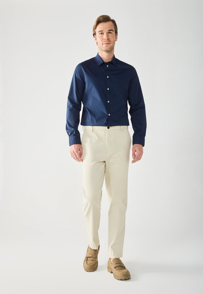 Marinefarbenes Hemd mit Knöpfen, cremefarbene Hose und braune Slipper. Das Hemd hat eine glatte Textur, während die Hose maßgeschneidert und leicht ist.