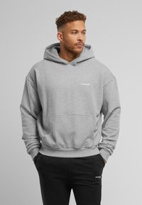 Lichtgrijze hoodie met een voorzak, ribbelboorden en een ribbelzoom. Bevat een witte tekstlogo op de borst. Gecombineerd met zwarte sweatpants.