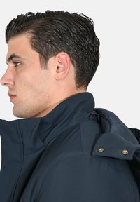 Giacca blu navy con colletto alto, superficie testurizzata, cerniera laterale e bottoni con dettagli dorati. Vista ravvicinata dei dettagli del colletto.