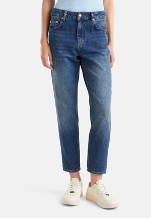 Femme portant un jean en denim bleu taille haute et des baskets blanches, debout devant un fond clair uni.