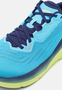 Chaussure de sport bleue avec tige en maille texturée, lacets bleu marine et semelle vert-jaune. Présente des superpositions lisses et des éléments de design en zigzag.