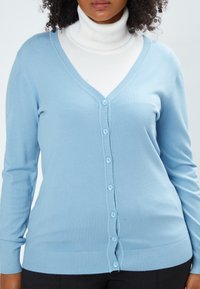 Hellblaues Cardigan aus weichem Material, mit V-Ausschnitt, durchgehenden Knöpfen und gerippten Bündchen sowie Saum. Getragen über einem weißen Rollkragenpullover.