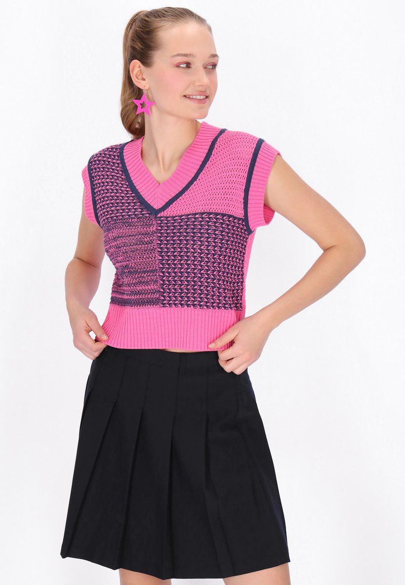 Kobieta ubrana w różowo-granatowy dziergany top bez rękawów, czarną plisowaną spódnicę i różowe kolczyki w kształcie gwiazdek, stojąca z rękami na biodrach.