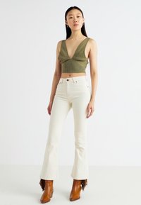 Olive green knit crop top with a deep V-neck and textured pattern, paired with white flared jeans and brown ankle boots with fringe accents.