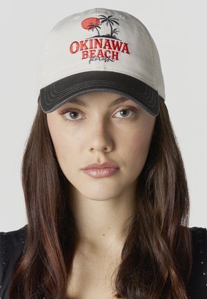 Joven con cabello largo y castaño que lleva una gorra beige y negra bordada con las palabras "Okinawa Beach" y gráficos de una palmera.