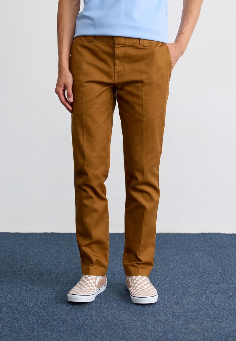 Personne portant un pantalon couleur rouille avec des plis marqués, associé à des chaussures slip-on à carreaux beiges et blanches sur un tapis bleu.