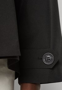 Mansetti musta takki, jossa on kuusikulmainen nappi, johon on kohokuvioitu "SAVE THE DUCK", sileä pinta ja räätälöity muotoilu.