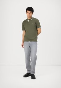 Olivovo zelená polo košeľa s bielymi detailmi, golierom, krátkymi rukávmi a logom. V kombinácii so svetlo šedými džínsy a čiernymi topánkami.