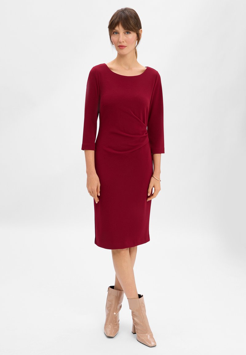 Abito bordeaux al ginocchio con maniche a tre quarti e dettaglio a torsione in vita; abbinato a stivali alla caviglia beige con punta quadrata.