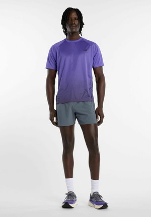 Mann steht in lila kurzärmeligem Hemd, grauer Shorts, weißen Socken und lila Laufschuhen vor einem schlichten weißen Hintergrund.