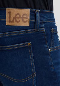 Nahaufnahme von dunkelblauen Jeans mit sichtbaren orangen Nähten und einem braunen Lederpatch mit der Aufschrift „Lee“ nahe dem Bund.