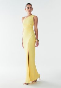 Gelbes, geripptes, einseitiges Abendkleid mit seitlichem Schlitz und figurbetonendem Design. Die Accessoires des Models umfassen ein goldenes Armband und Riemchen-High Heels.