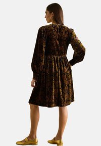 Samtkleid mit Blumendruck in dunklen Farben, geraffter Taille und langen Ärmeln. Kombiniert mit goldenen flachen Schuhen.