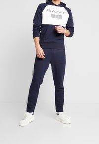 Sudadera navy con el frente blanco, con el logo de GANT y bolsillo tipo canguro. Combinada con pantalones de chándal navy y zapatillas blancas con detalles azules.