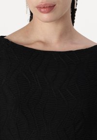 Vestido preto com tecido texturizado apresentando um padrão ondulado. O decote é amplo e o visual é completado com um detalhe de brincos perolados.