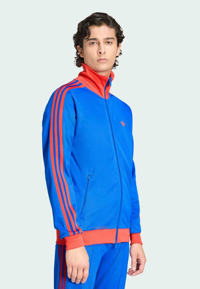 Young man wearing a blue and red zip-up athletic jacket with matching pants, standing against a plain light background.