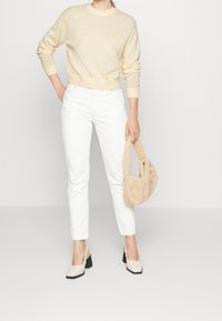 Femme portant un pull crème et un pantalon blanc, tenant un sac à main en fourrure beige et portant des chaussures à talons bloc crème avec une bride derri�ère.