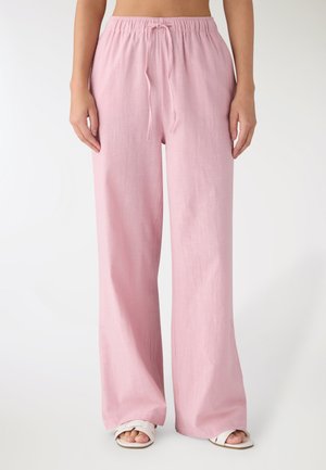 Donna che indossa pantaloni larghi di lino rosa chiaro con vita elastica e coulisse, abbinati a sandali bianchi aperti sulle dita.