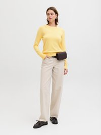 Vrouw draagt een gele trui, beige wijde broek, zwarte riemtas en zwarte schoenen, staand tegen een effen lichte achtergrond.