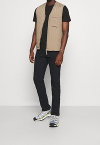 Chaleco acolchado beige con cremallera frontal y un bolsillo, usado sobre una camiseta negra, combinado con pantalones negros y zapatillas deportivas grises.
