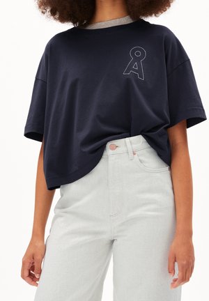 Laivansinis ylisuuri t-paita, harmaalla sisäkauluksella, jossa on valkoisella rajattu "A"-muotoilu vasemmassa rinnassa, yhdistettynä vaaleisiin farkkuihin.