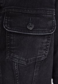 Μαύρο τζάκετ από τζιν με τσέπη στο στήθος και μαύρο κουμπί με κόψιμο, εμφανείς λεπτομέρειες ραφής και υφή υφάσματος.