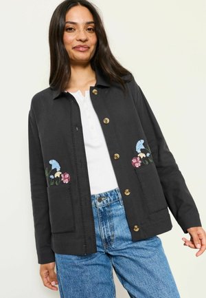 Frau trägt eine schwarze, geknöpfte Jacke mit Blumenstickerei auf den Taschen, ein weißes Hemd und blaue Jeans, steht vor einem schlichten hellen Hintergrund.