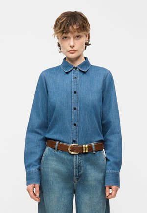 Osoba ubrana w zapinaną na guziki niebieską koszulę jeansową wpuszczoną w wysokowodne niebieskie dżinsy z brązowym skórzanym paskiem na prostym tle.