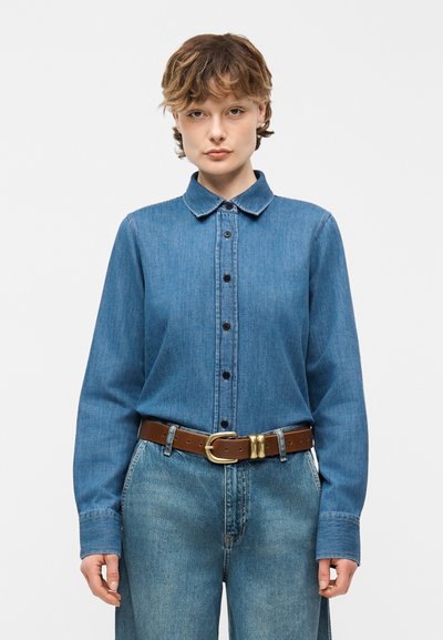 Personne portant une chemise en denim bleue boutonnée rentrée dans un jean taille haute bleu avec une ceinture en cuir marron sur un fond uni.