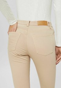 Béžové skinny džíny s hladkým povrchem, které mají dvě zadní kapsy a hnědou koženou nášivku na pase.