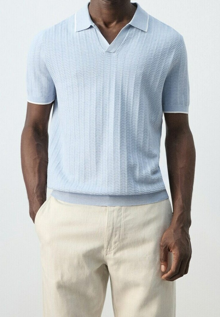 Homme portant un polo en maille bleu clair à manches courtes avec un motif texturé et un pantalon beige, debout, une main dans la poche.