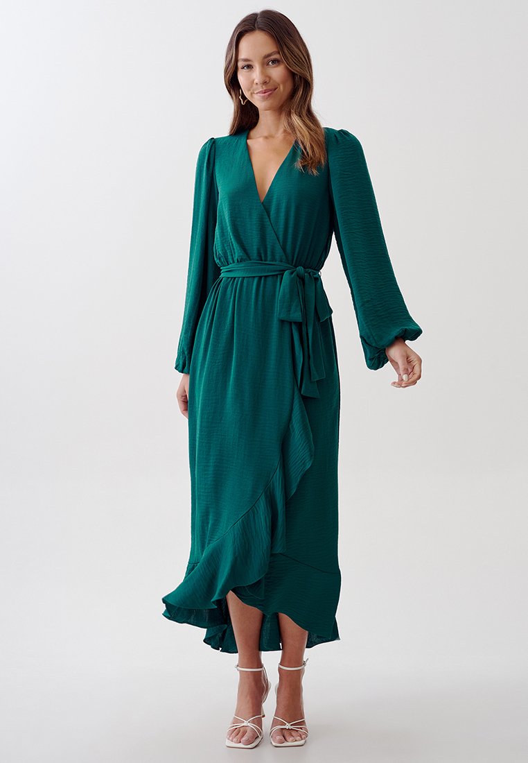 Robe enroulée verte avec des manches longues, en tissu texturé, un lien à la taille et un ourlet volanté, associée à des sandales blanches à lanières.